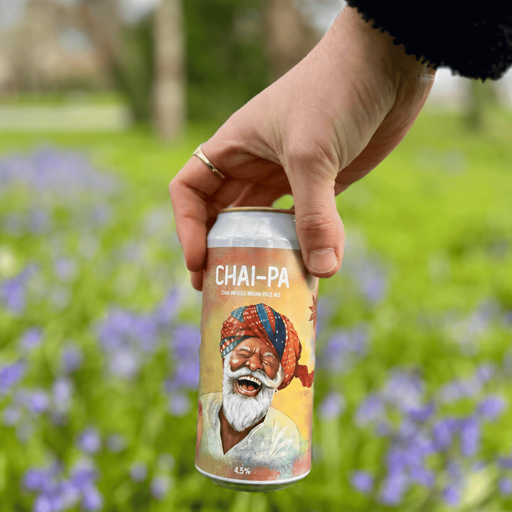 Hand holding a can of 'Chai-Pa' beer with a colorful label featuring an illustration of a man in a turban against a blurred natural background.