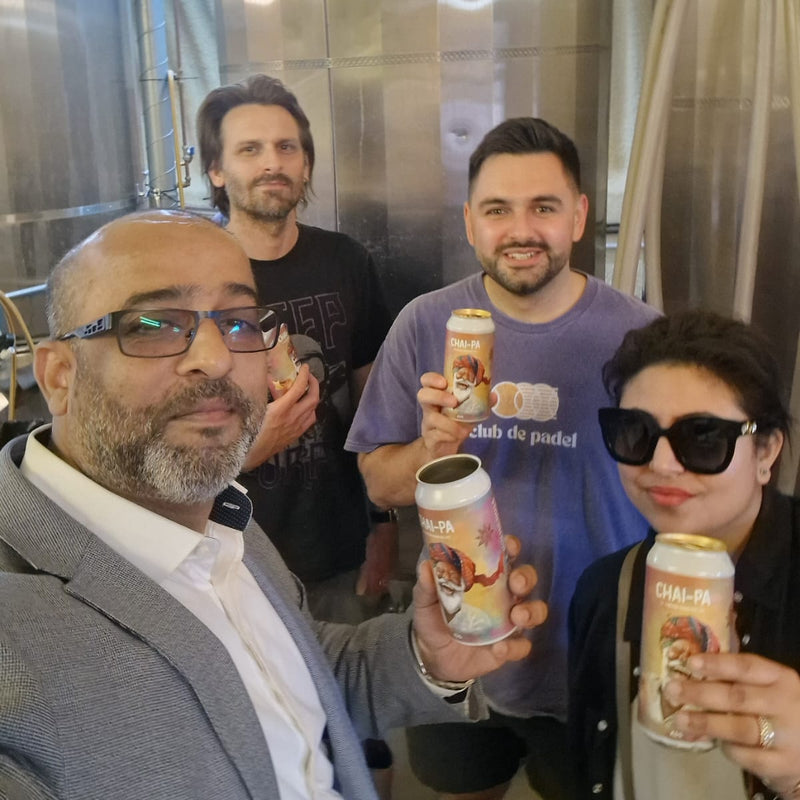 Four people holding cans in front of a large metal tank, likely in a brewery setting.